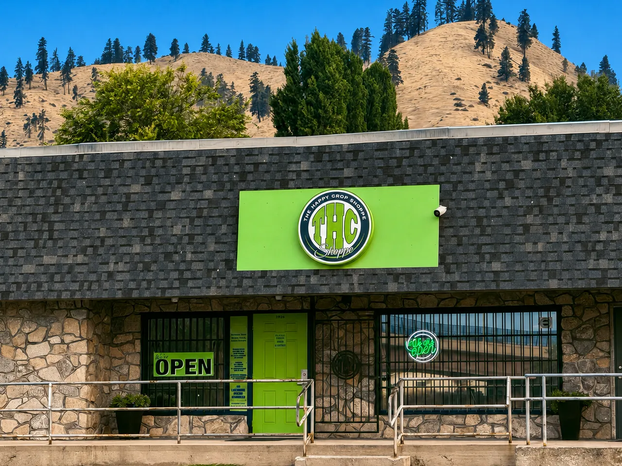 The Happy Crop Shoppe in Cashmere: a stone fronted shoppe under a dark shingle roof, a bright green THC Shoppe sign board over the door, OPEN signs in the windows and pine covered hills behind.
