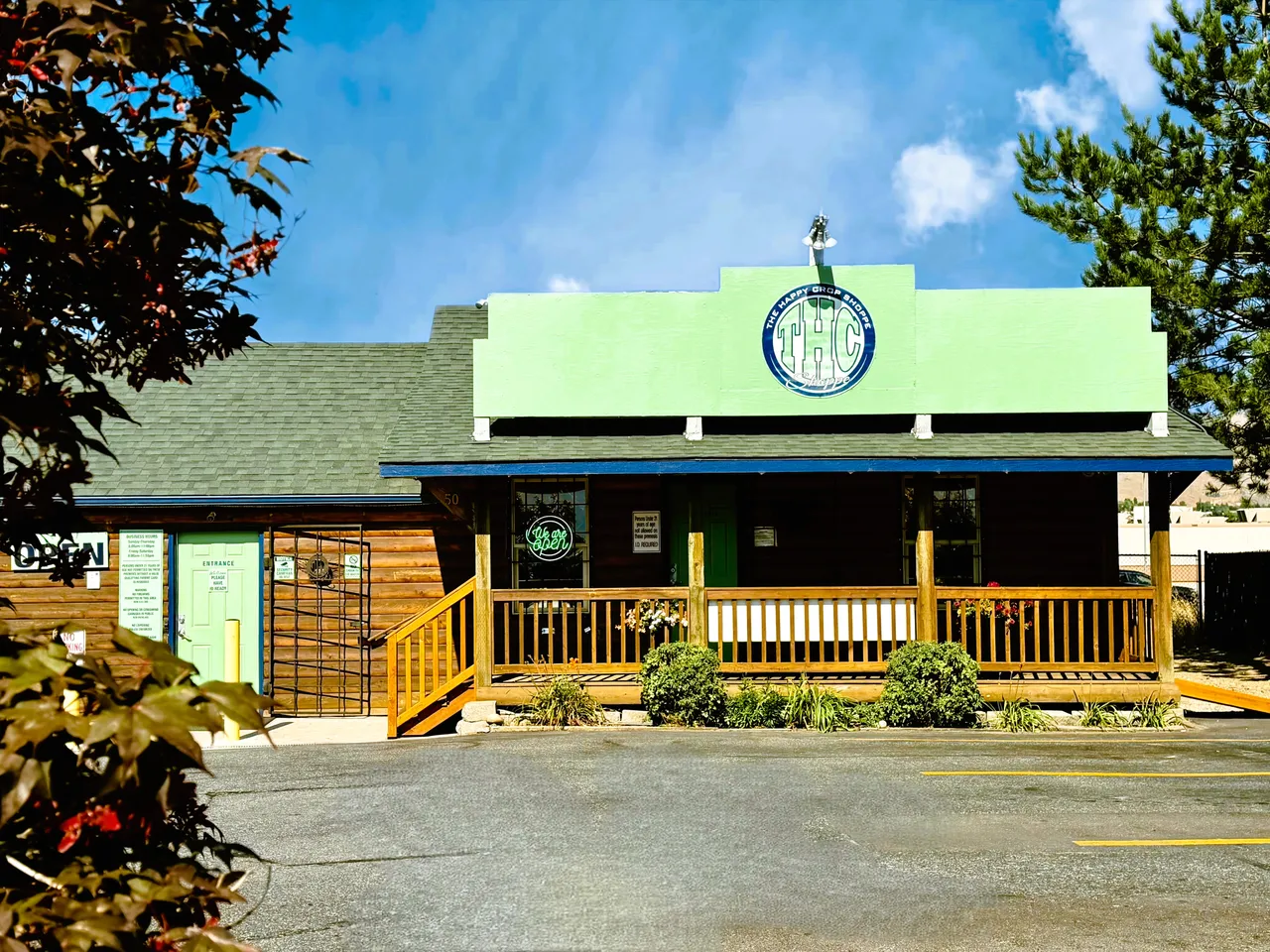 The Happy Crop Shoppe in East Wenatchee: a log building with a covered porch and wooden railings, a pale green sign board carrying the THC Shoppe roundel above it, pines and a blue sky behind.