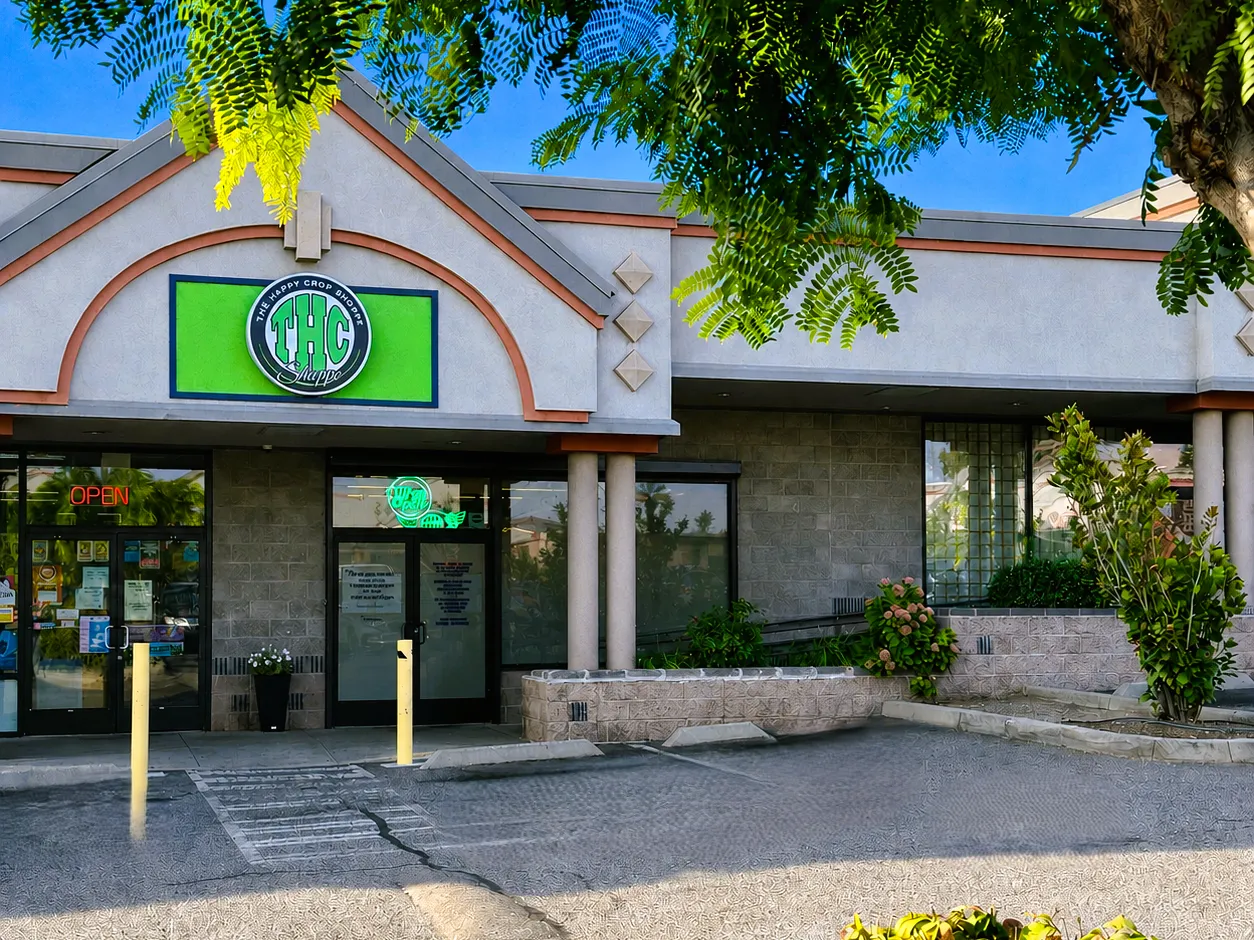 The Happy Crop Shoppe in Wenatchee: a stucco strip mall front under a peaked gable, the green THC Shoppe sign board beneath the arch, an OPEN sign in the window and a leafy tree over the parking lot.